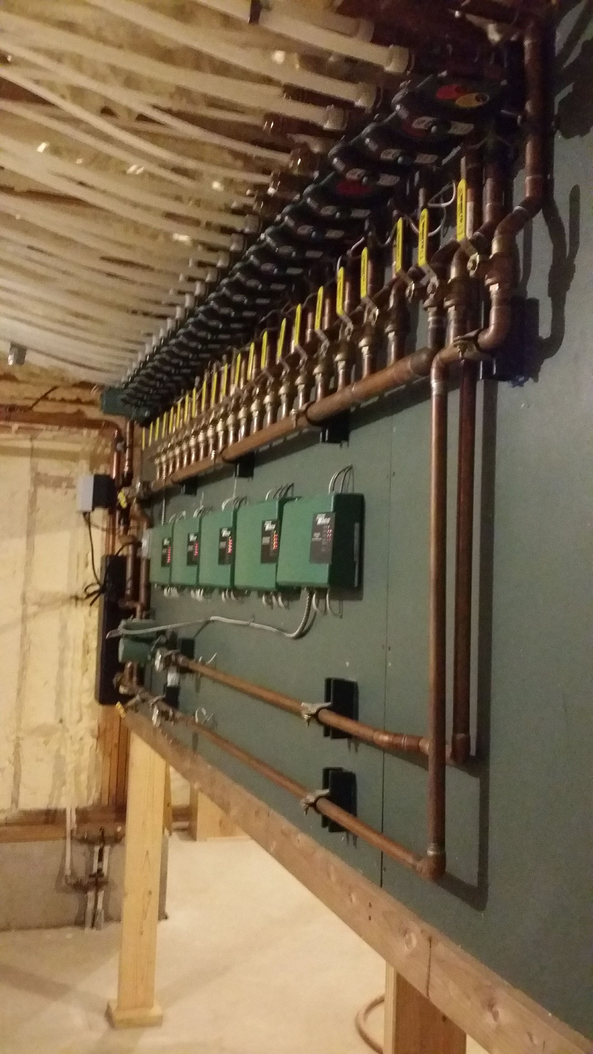 Copper pipes, valves, and green electrical components mounted on a wooden framework in a basement.