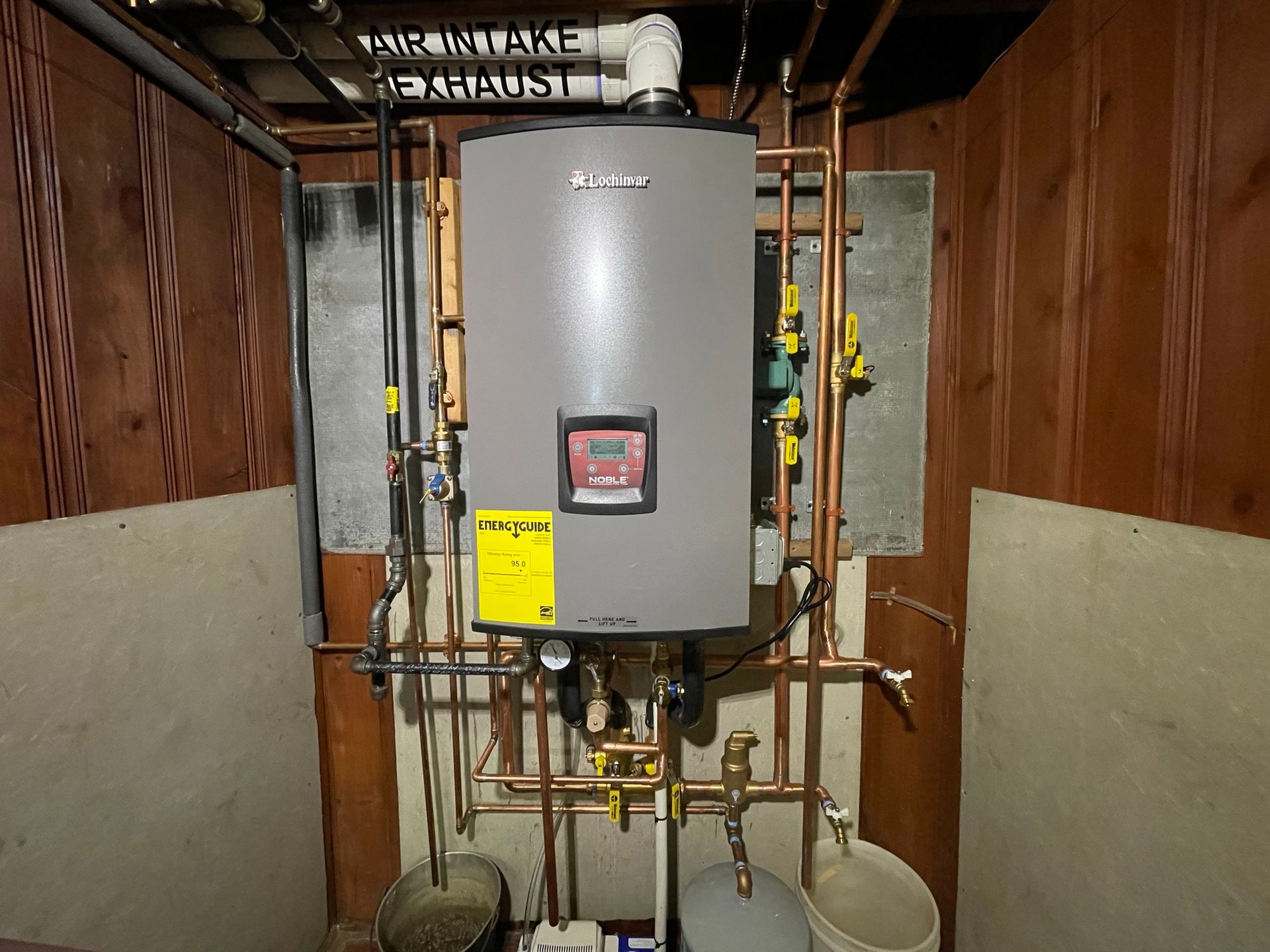 A wall-mounted gray gas boiler with copper pipes, installed in a utility room with wooden paneling.