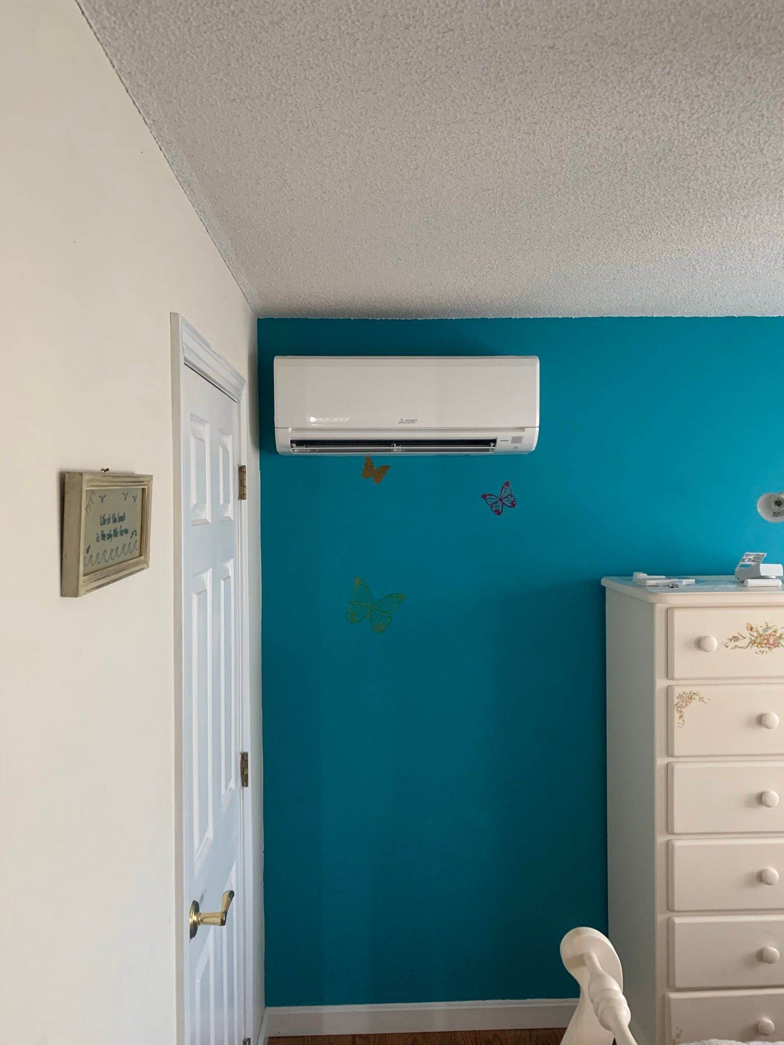 White air conditioner on a teal wall in a bedroom with a white dresser.