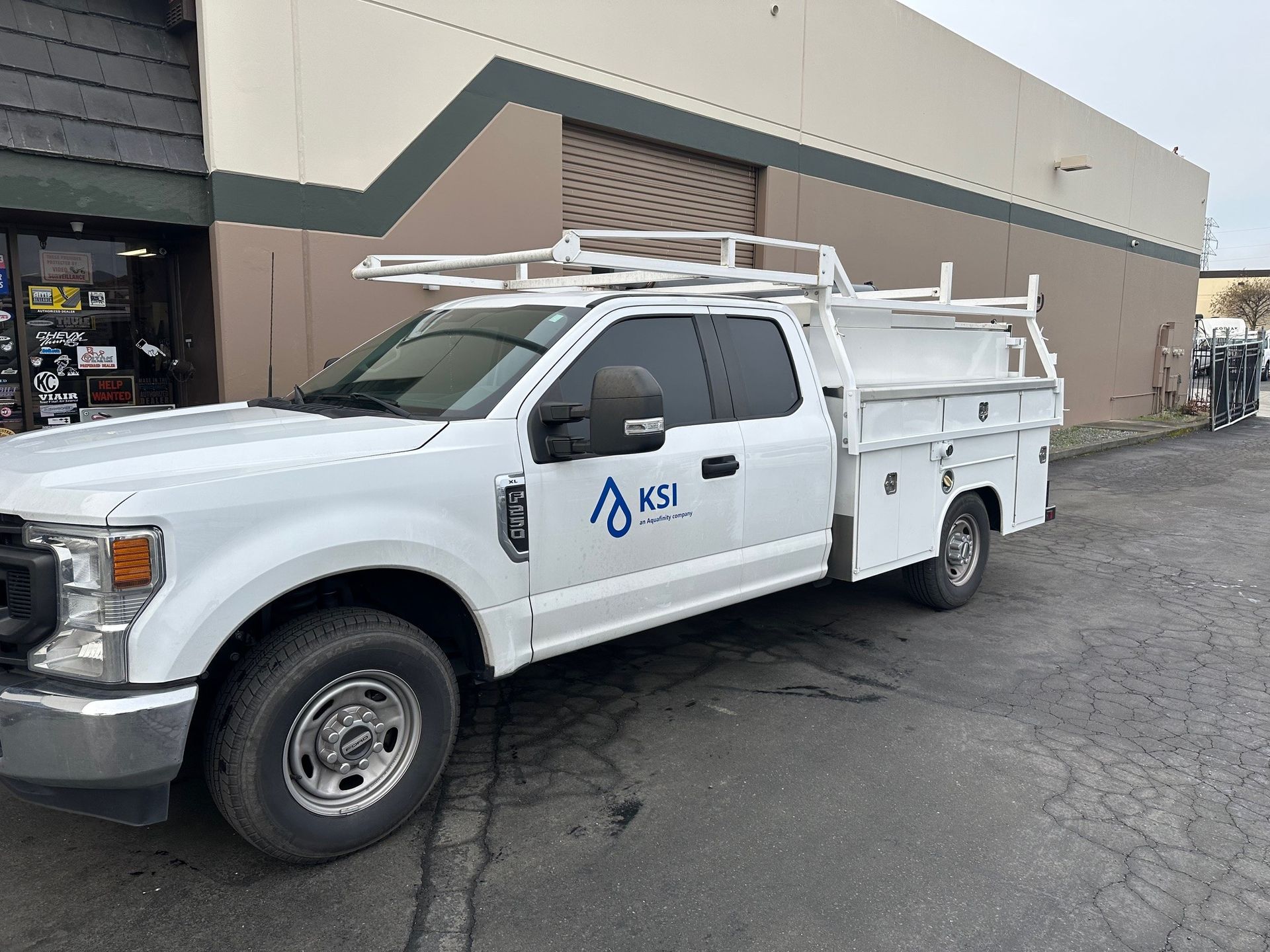 A white truck with a ladder rack on the roof is parked in front of a building.