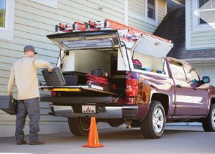 Man unloading gear from a maroon pickup truck with open canopy in a driveway, orange cone behind it
