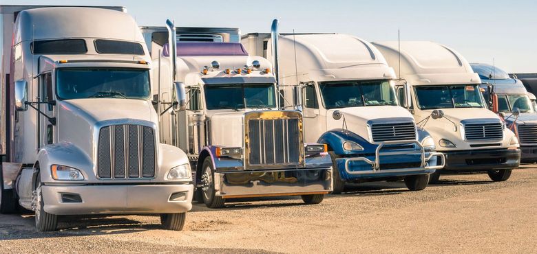 A row of semi trucks are parked in a dirt lot.