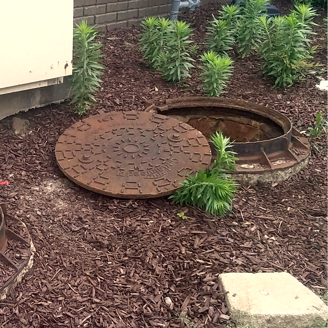 A rusty manhole cover is sitting in the dirt next to a brick building.