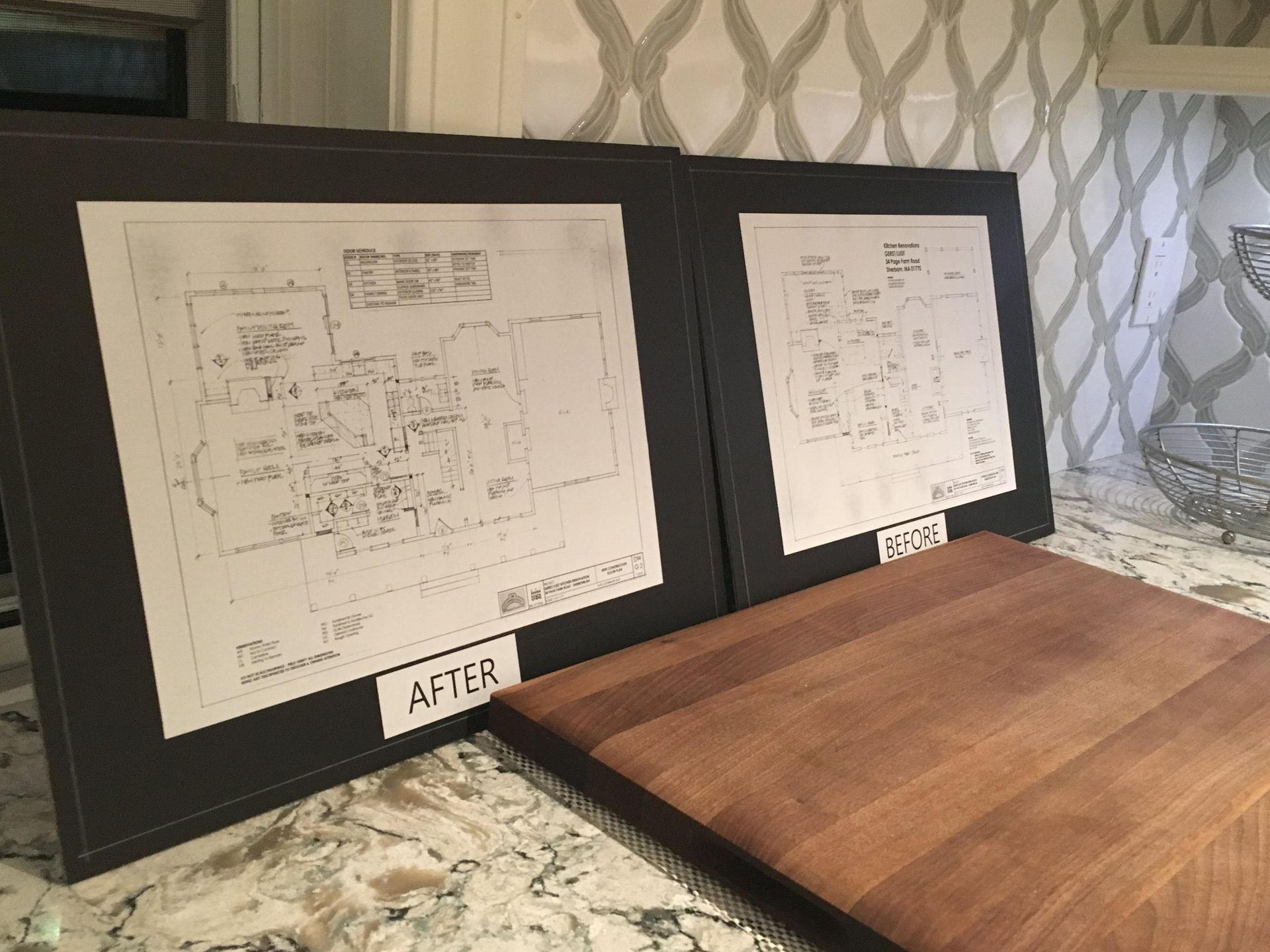Two framed drawings of a house are on a cutting board on a counter.