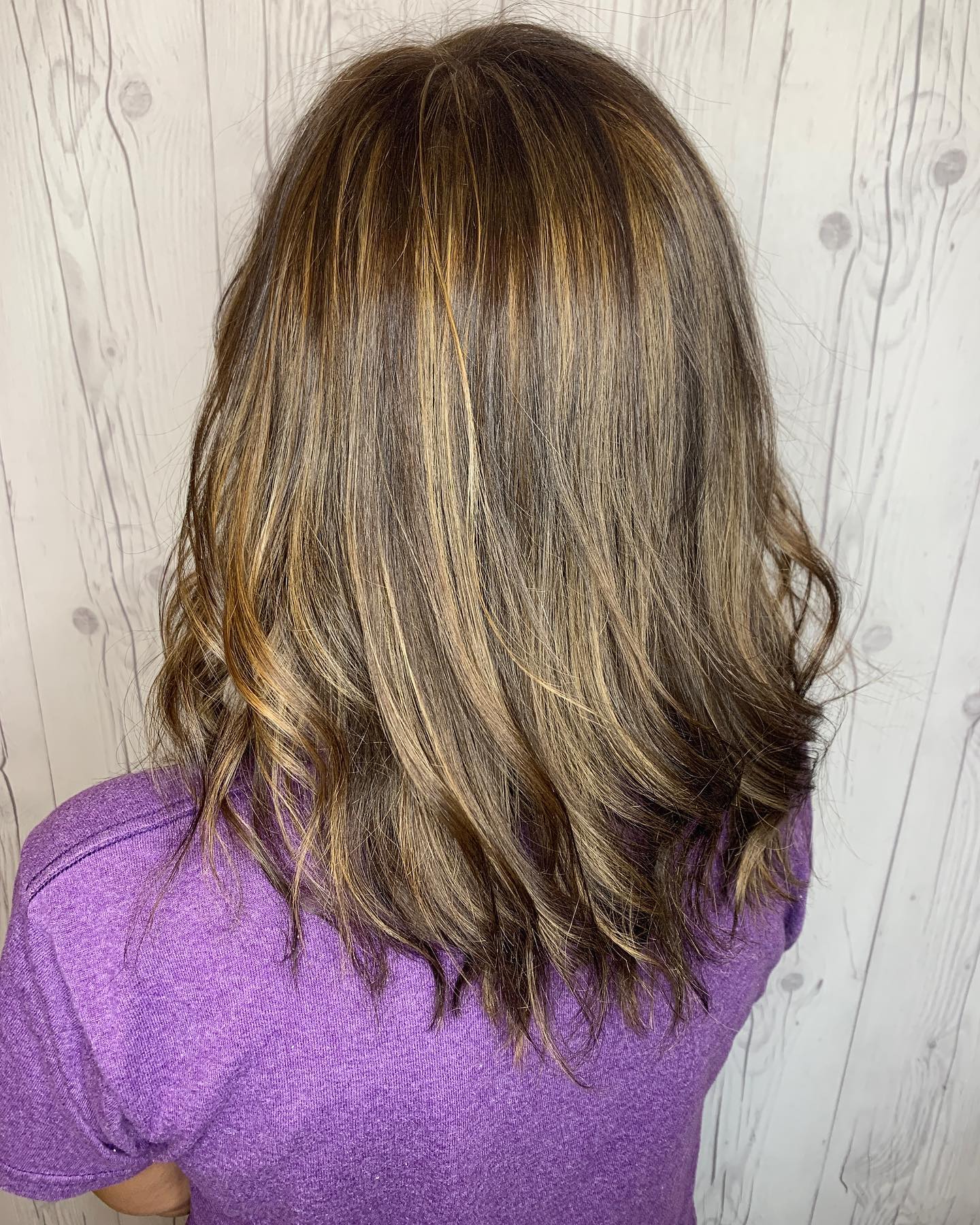 A woman with long brown hair is wearing a purple shirt and standing in front of a wooden wall.