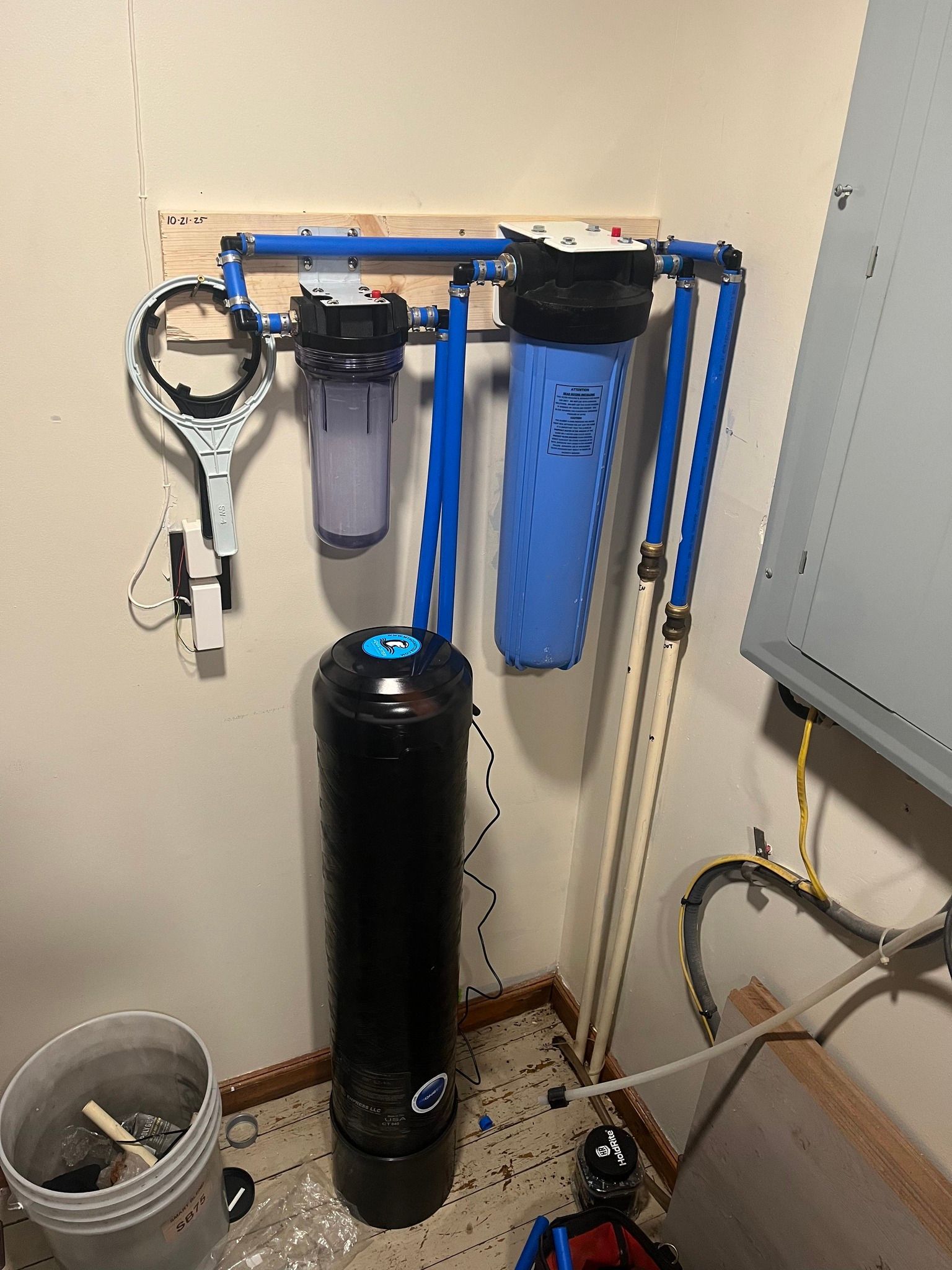 Water filtration system with three filters and a softener, mounted on a wall with blue pipes.