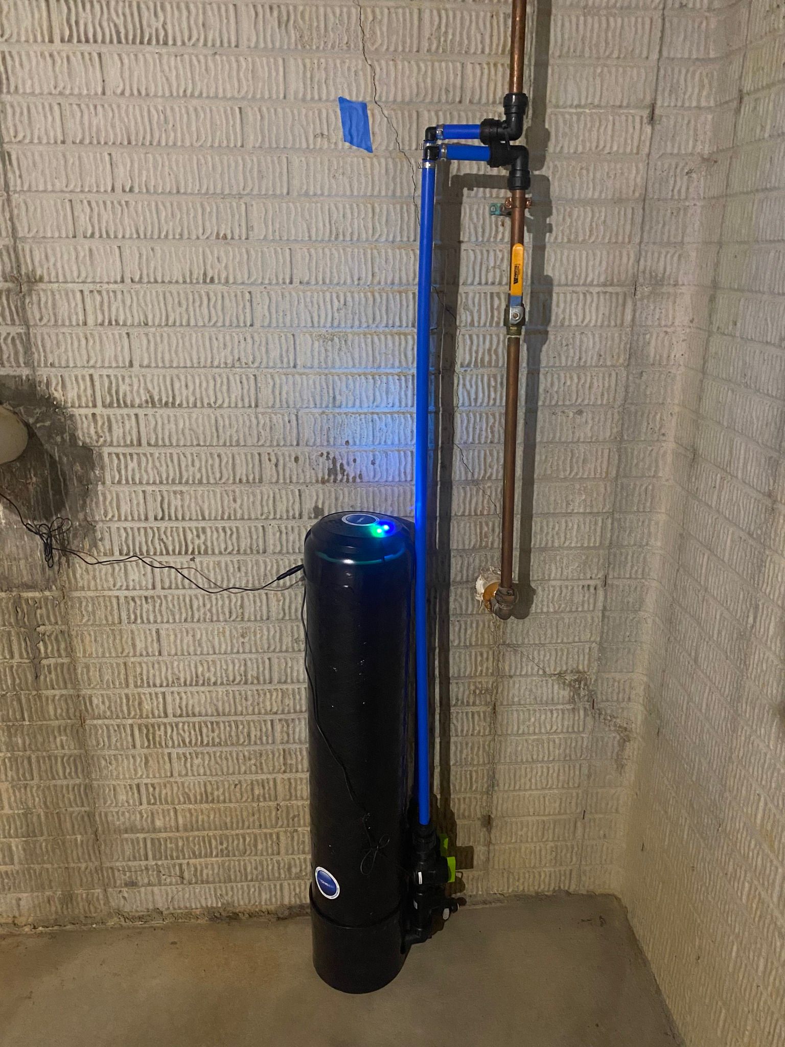 Black water softener against a cinder block wall, connected to copper pipes and a blue bypass.
