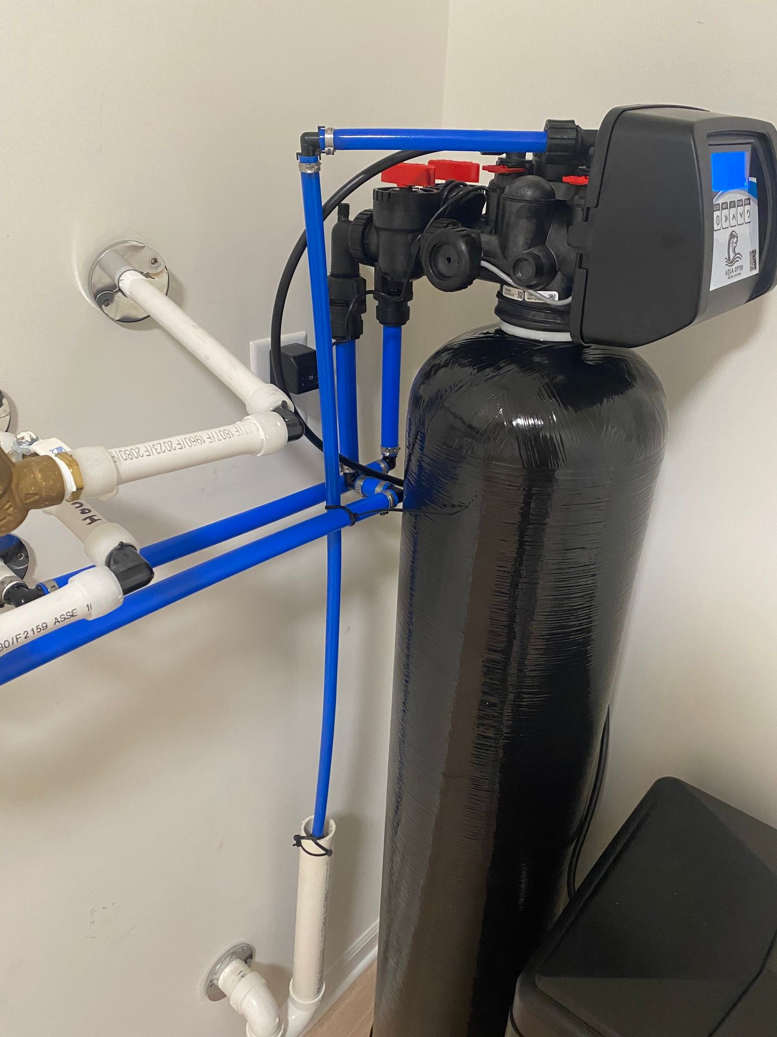 Water softener system with black tank, blue pipes, and control panel on a white wall.
