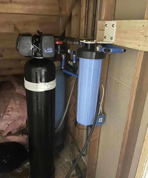Water filtration system: black tank, blue filter, pipes, and control panel against wood paneling.
