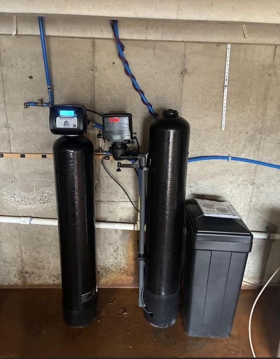 Two black water filtration tanks with control panels, connected by blue piping, mounted on a concrete wall.