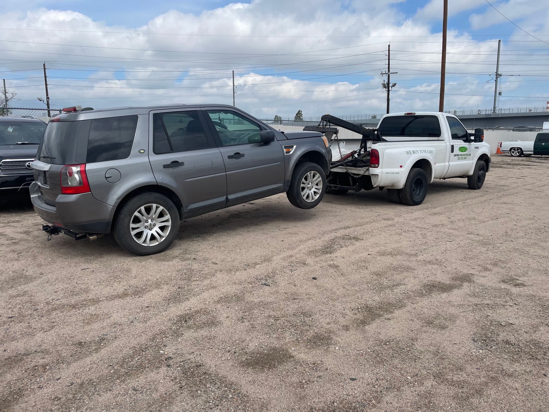 Gray SUV being towed by a white tow truck in a gravel lot under a cloudy sky