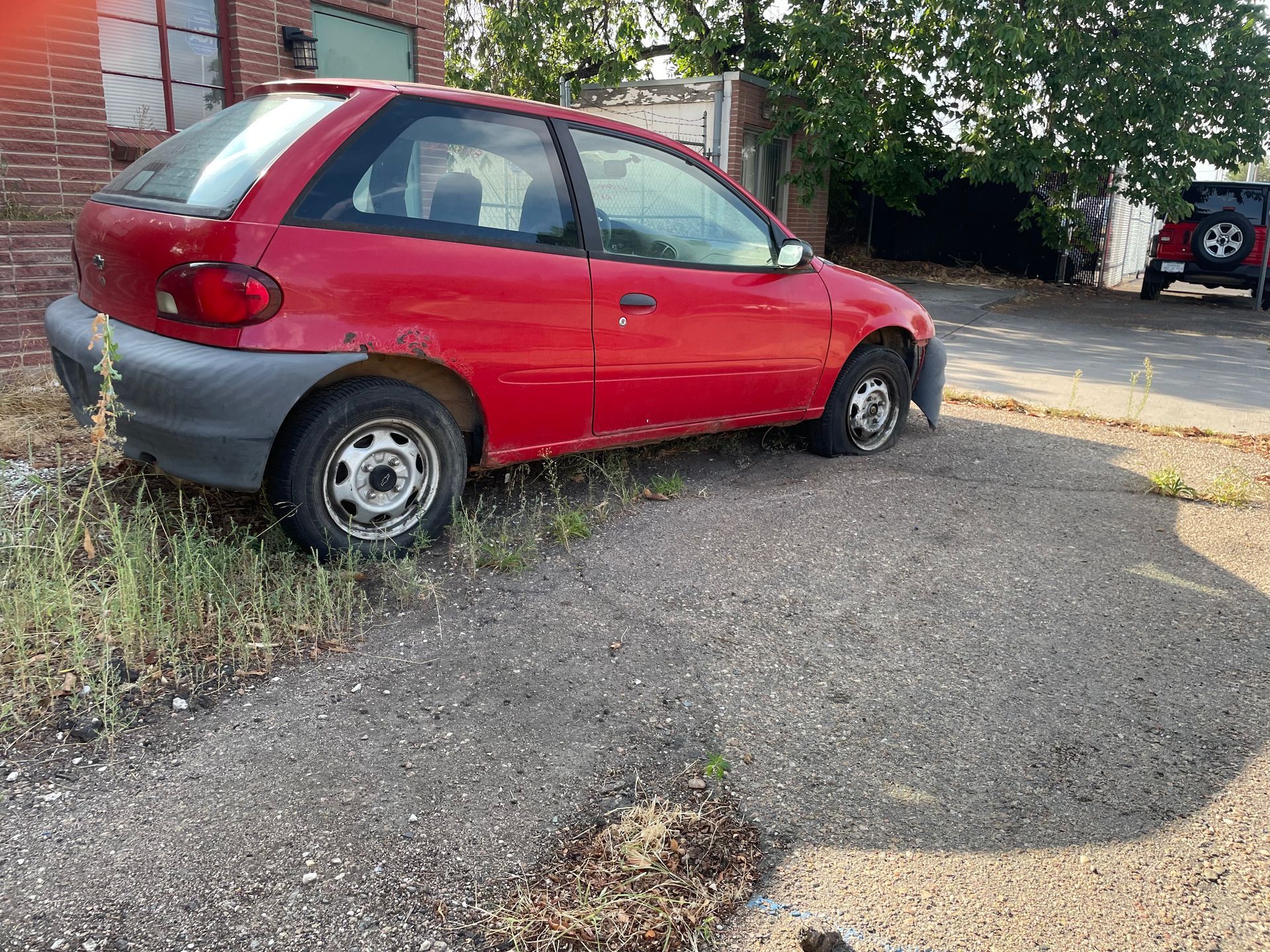 Red hatchback parked on an overgrown gravel lot beside a brick building.