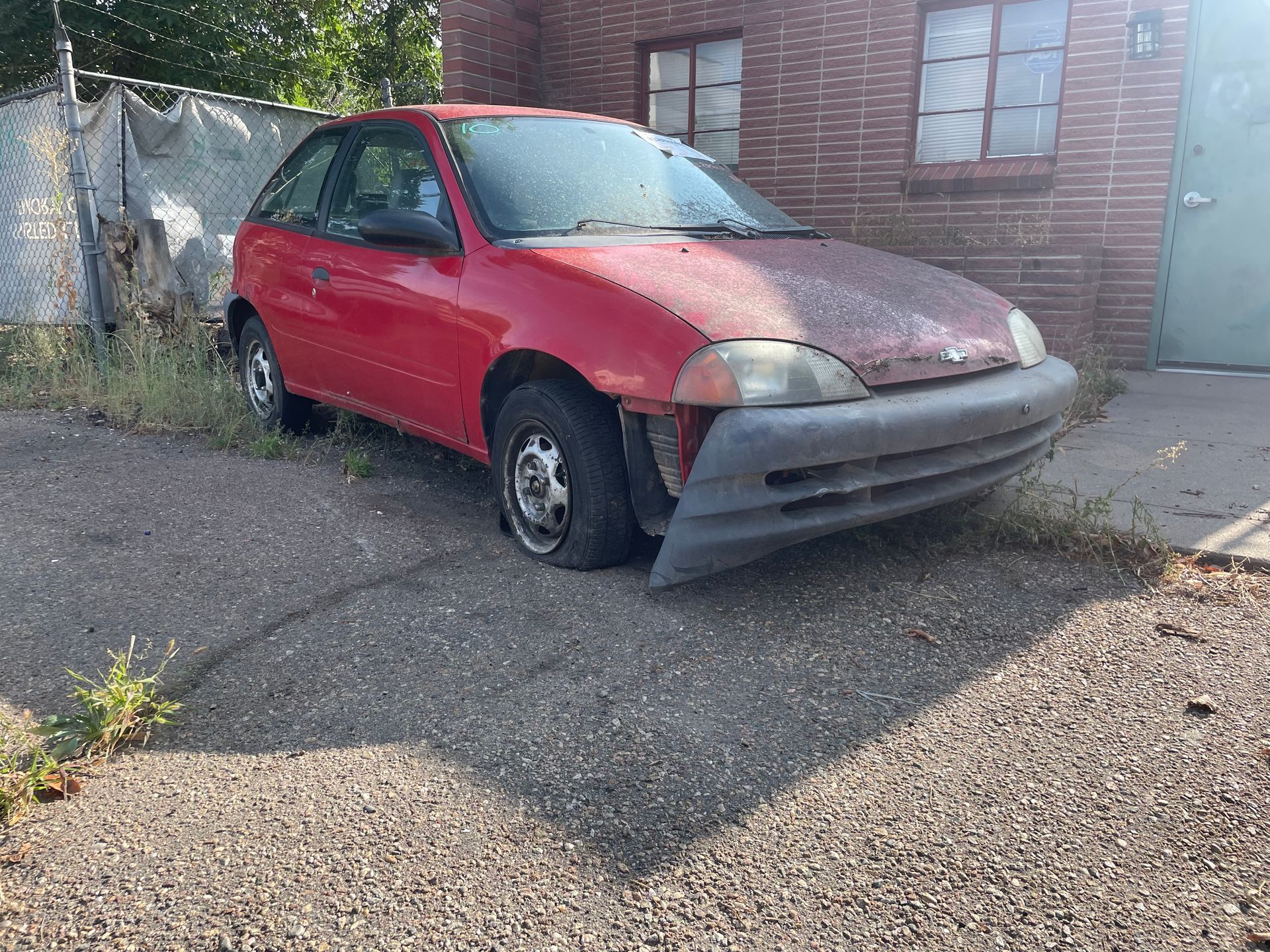 Red compact car with front-end damage parked on gravel beside a building and gate.