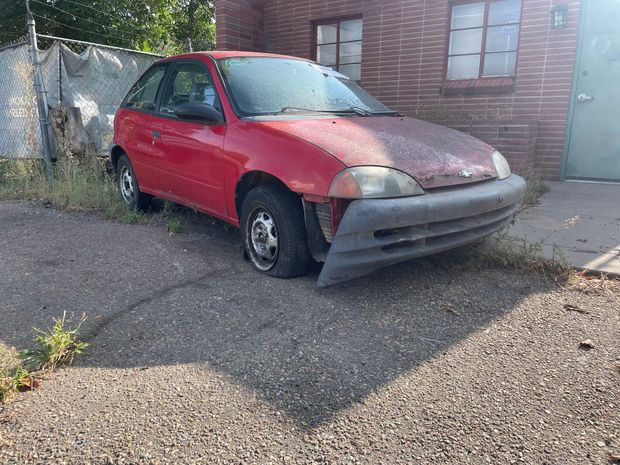 Red compact car with front-end damage parked on gravel beside a building and gate.