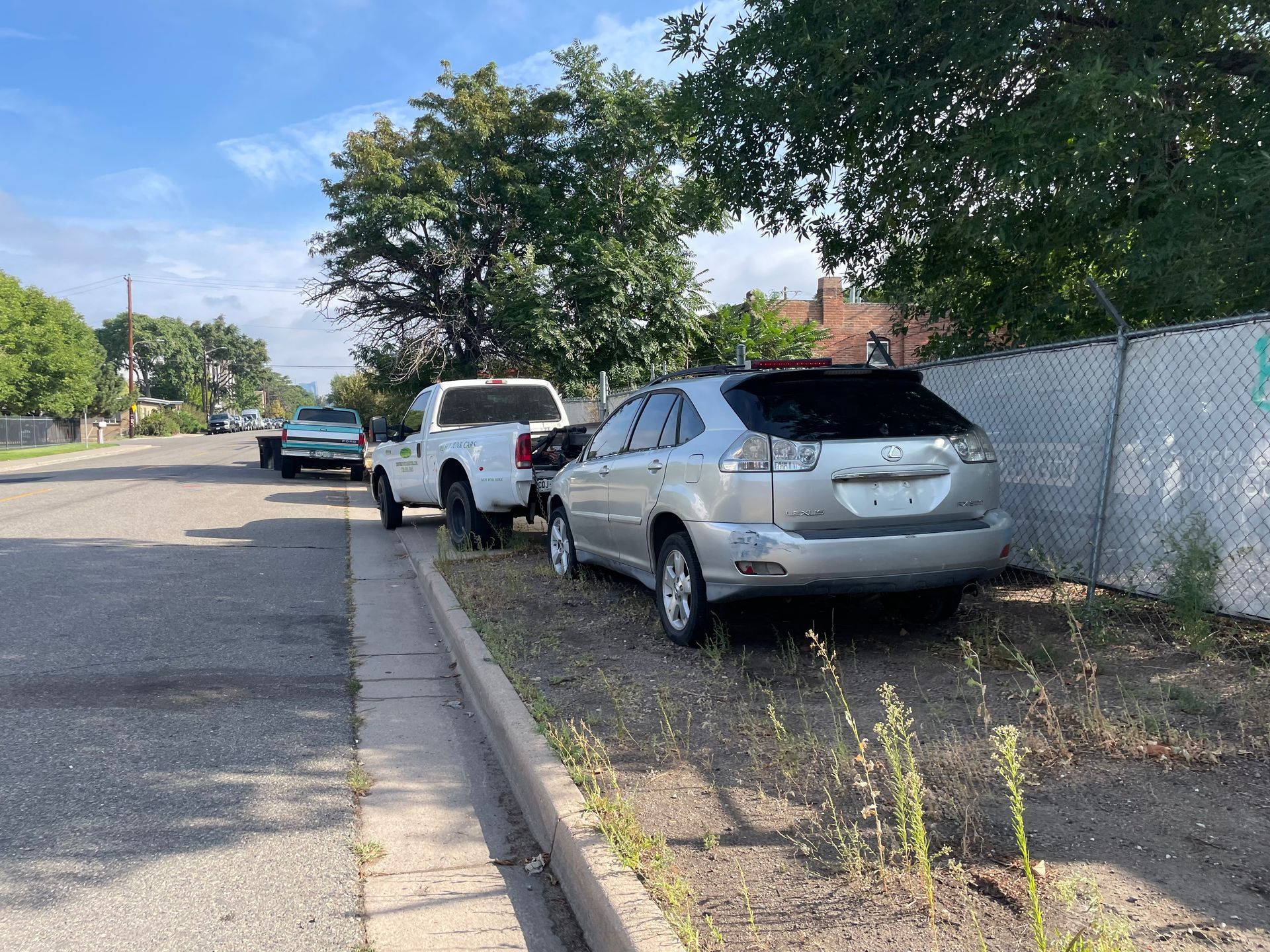 Parked cars along a curb beside a chain-link fence on a sunny residential street