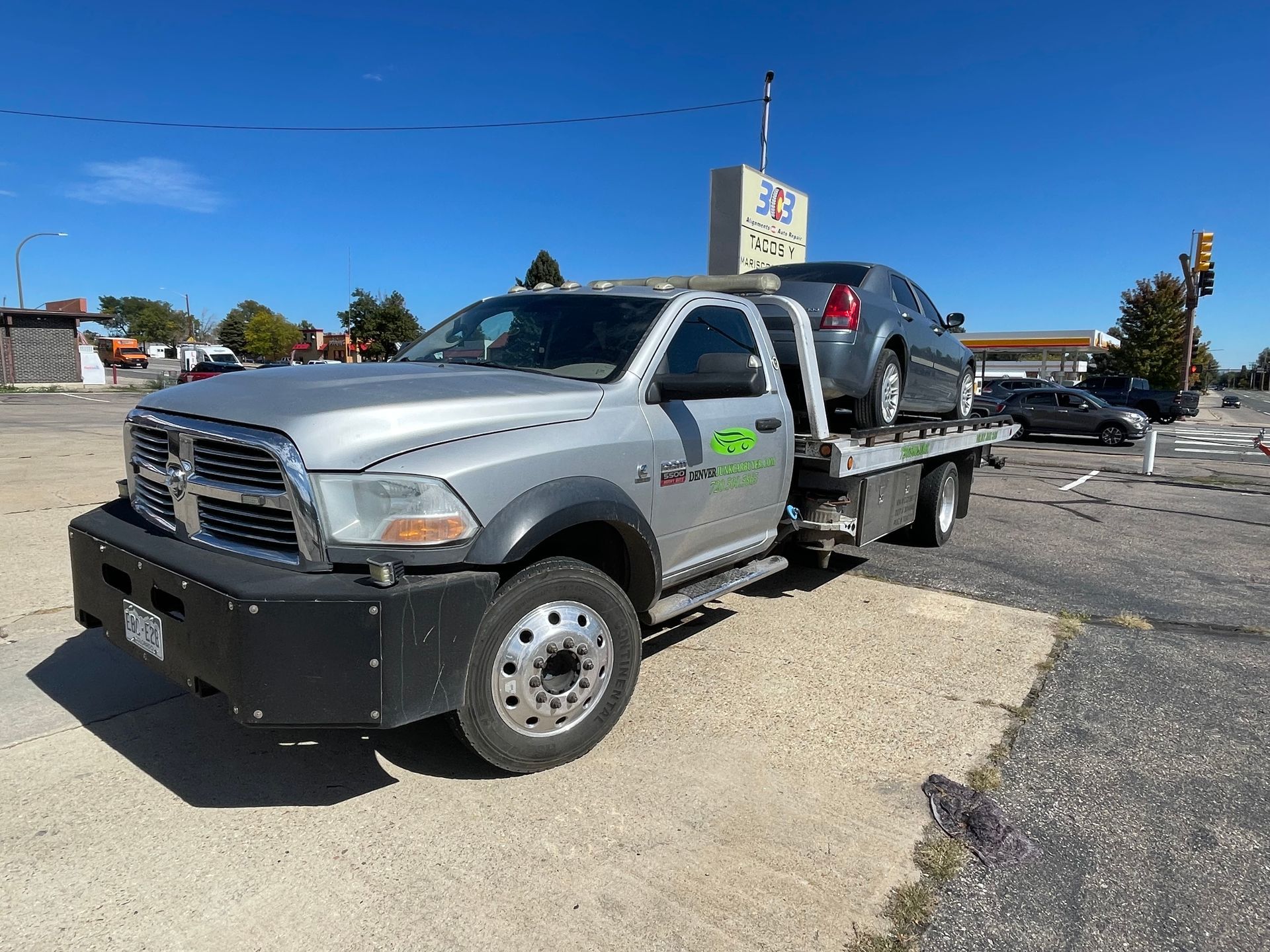 Silver tow truck carrying a black SUV on a flatbed in a parking lot under a clear blue sky