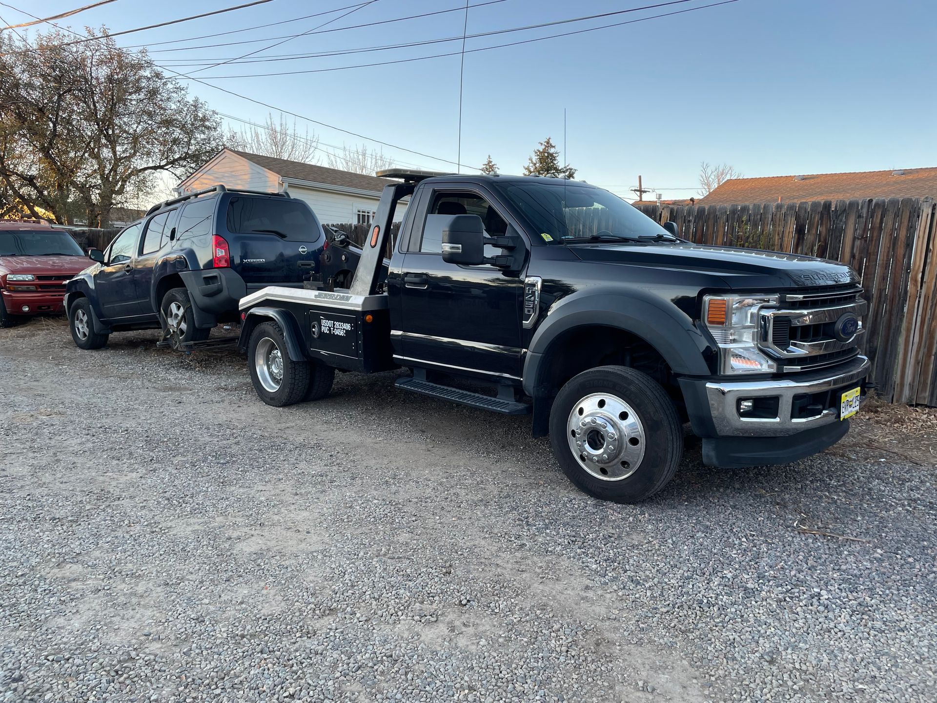 Black flatbed tow truck parked on a gravel lot, with a black SUV behind it.