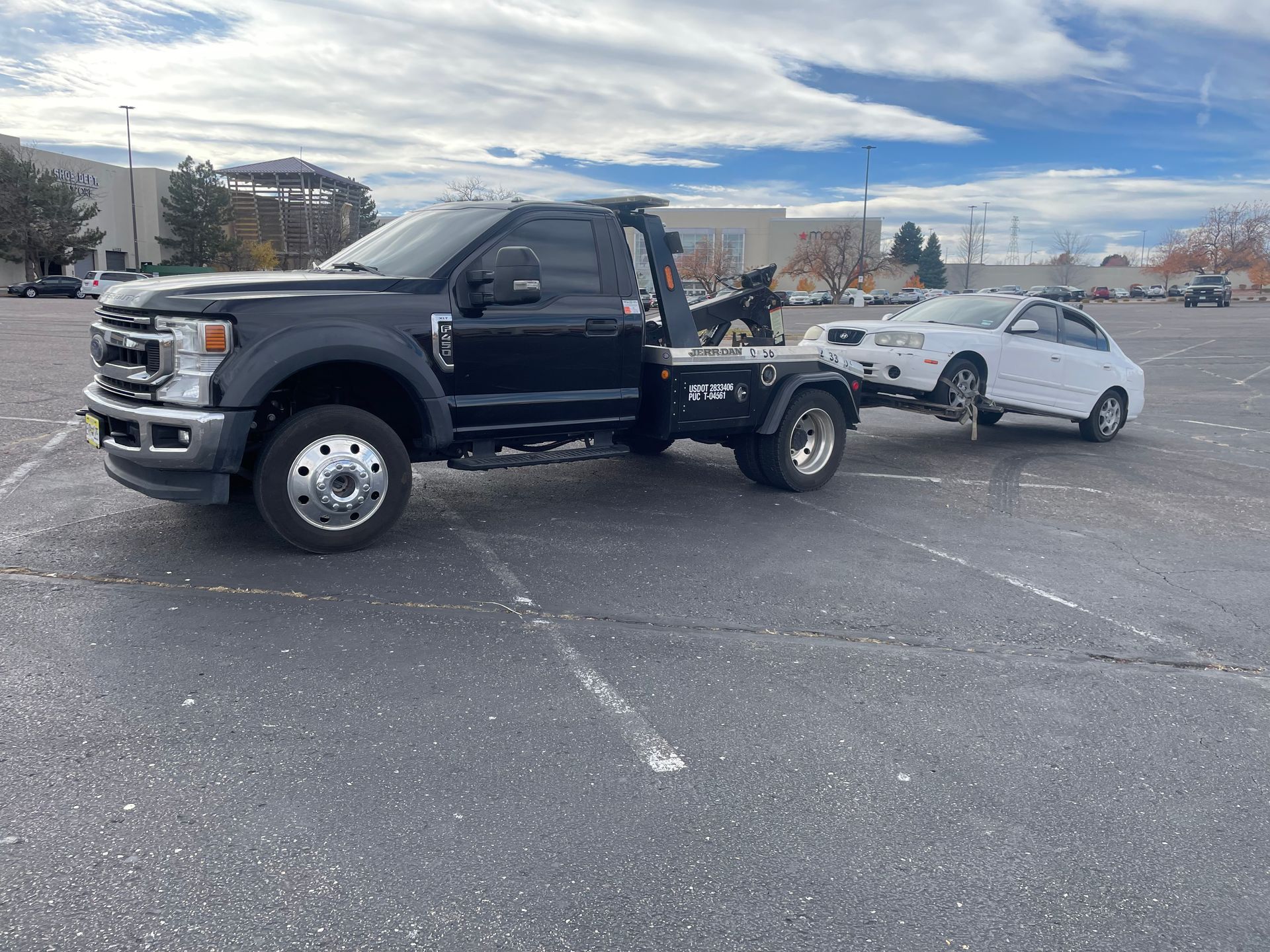 Black flatbed tow truck hauling a white sedan in a parking lot under a cloudy sky