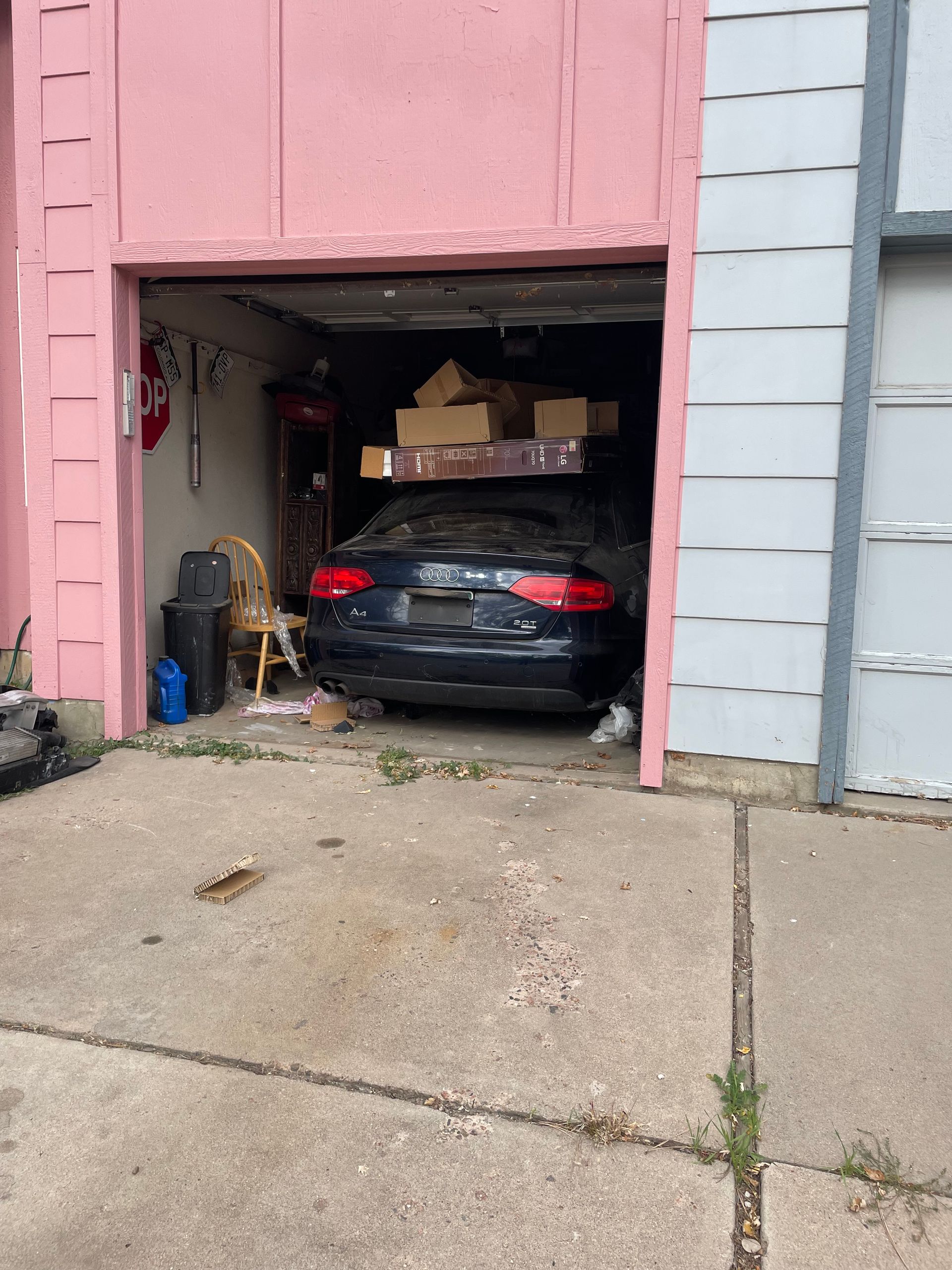 Black sedan parked in a cluttered garage with the door open, pink and gray exterior visible.