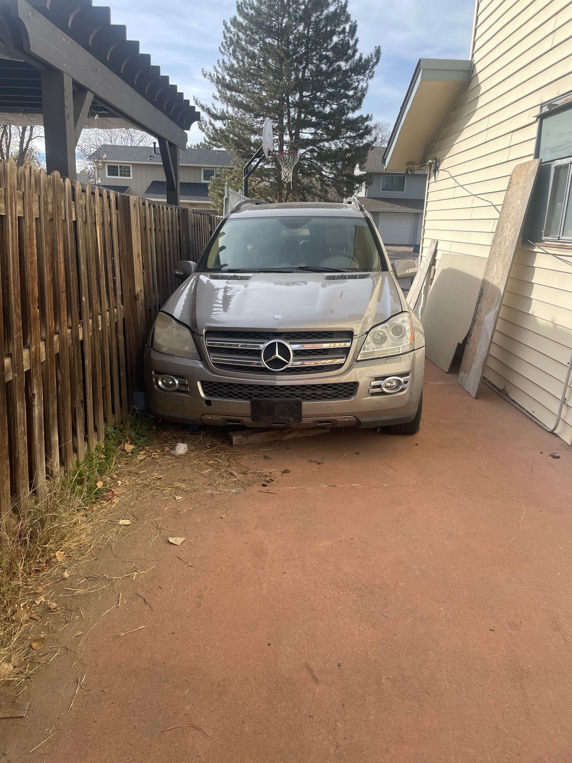 Silver Mercedes SUV parked in a narrow dirt driveway beside a wooden fence and house wall