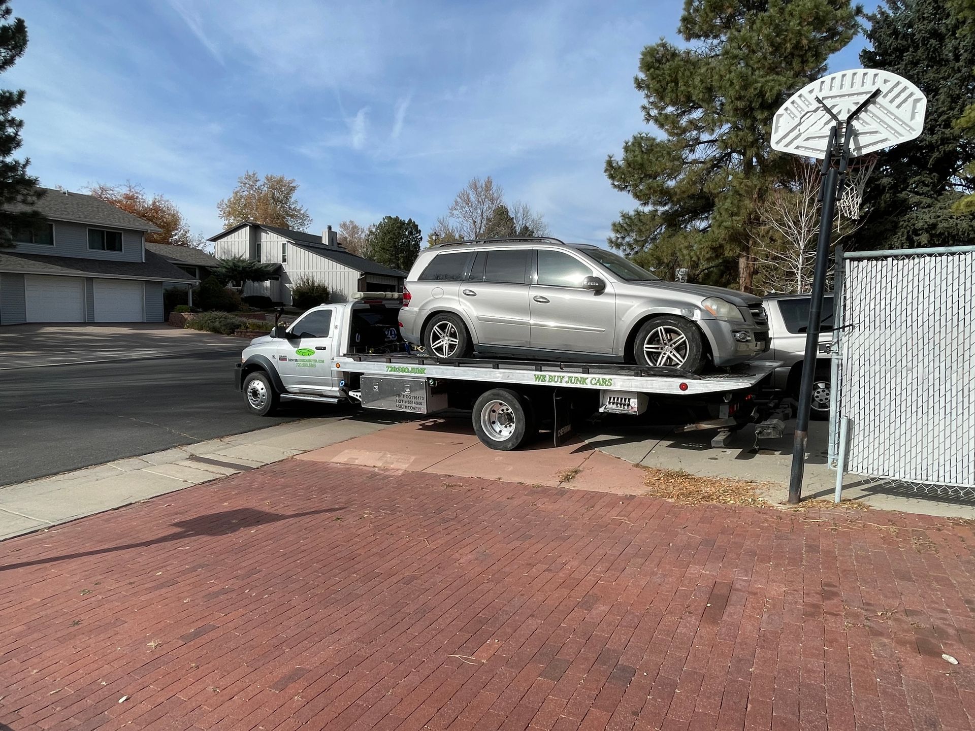 Tow truck loading a silver car on a flatbed in a suburban driveway, with a basketball hoop nearby