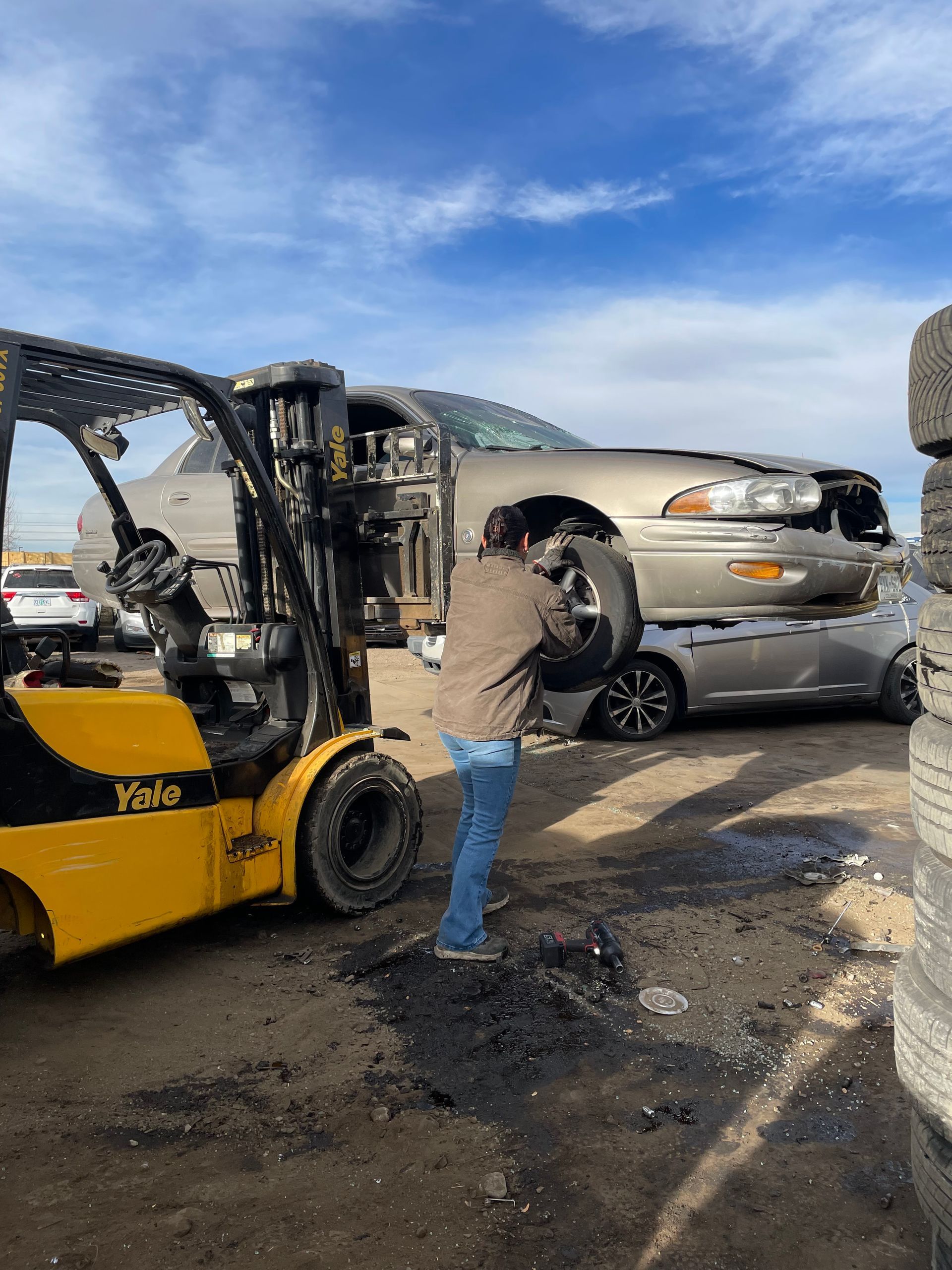 Worker inspecting rear damage on a silver car beside a yellow forklift at a roadside accident scene