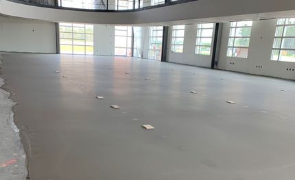 A wide, empty room with a smooth gray concrete floor and large windows along the far wall under construction.