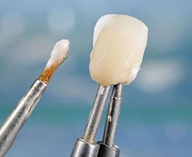 Dental tools holding a white tooth crown and a small brush against a blue background.