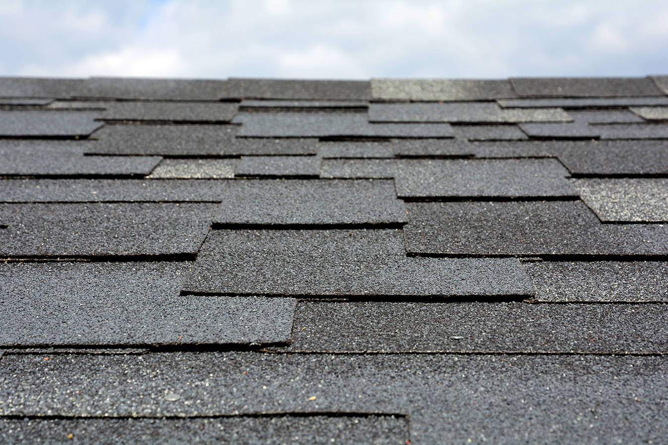 Gray asphalt shingles on a rooftop, textured and overlapping, against a cloudy sky.