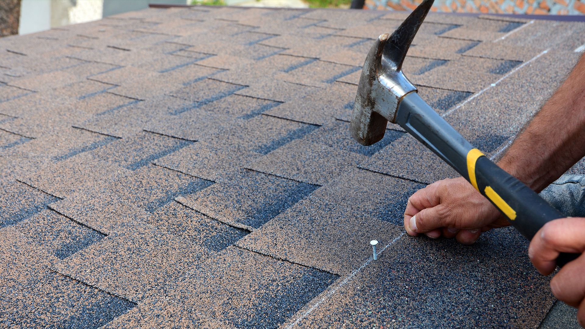 A person hammering a nail into asphalt roof shingles.