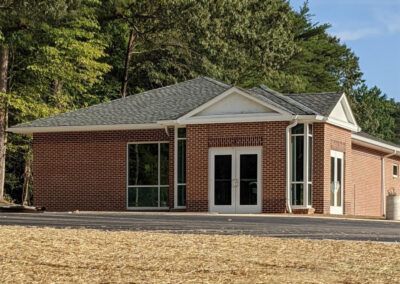 Brick building with large windows and a gray roof, surrounded by trees and a paved area.