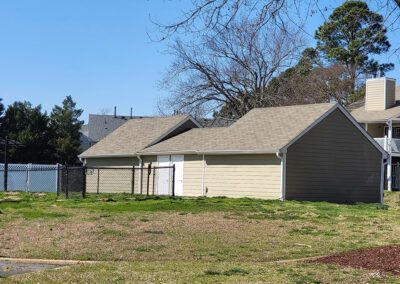 A light brown building with tan siding and a dark roof behind a fenced grassy area on a sunny day.