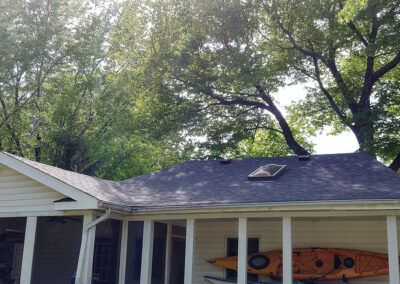 House with a dark shingled roof, skylight, and porch; trees in the background, a yellow kayak mounted on the wall.