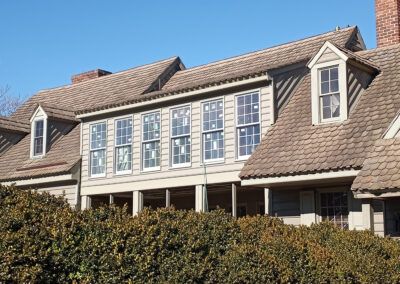 Tan-colored house with multiple windows and a brown shingle roof, obscured by green bushes.