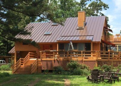 Brown wooden house with a metal roof, deck, and chimney in a grassy setting.