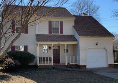 Beige two-story house with red shutters and door, white garage door, brown roof, and bare tree in front.