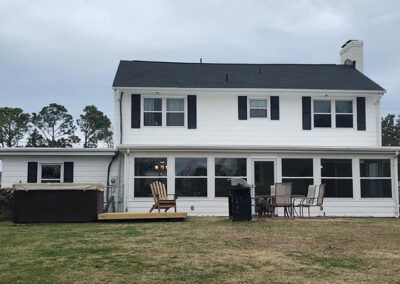 White two-story house with black shutters, a sunroom, and a hot tub on a grassy lawn under an overcast sky.