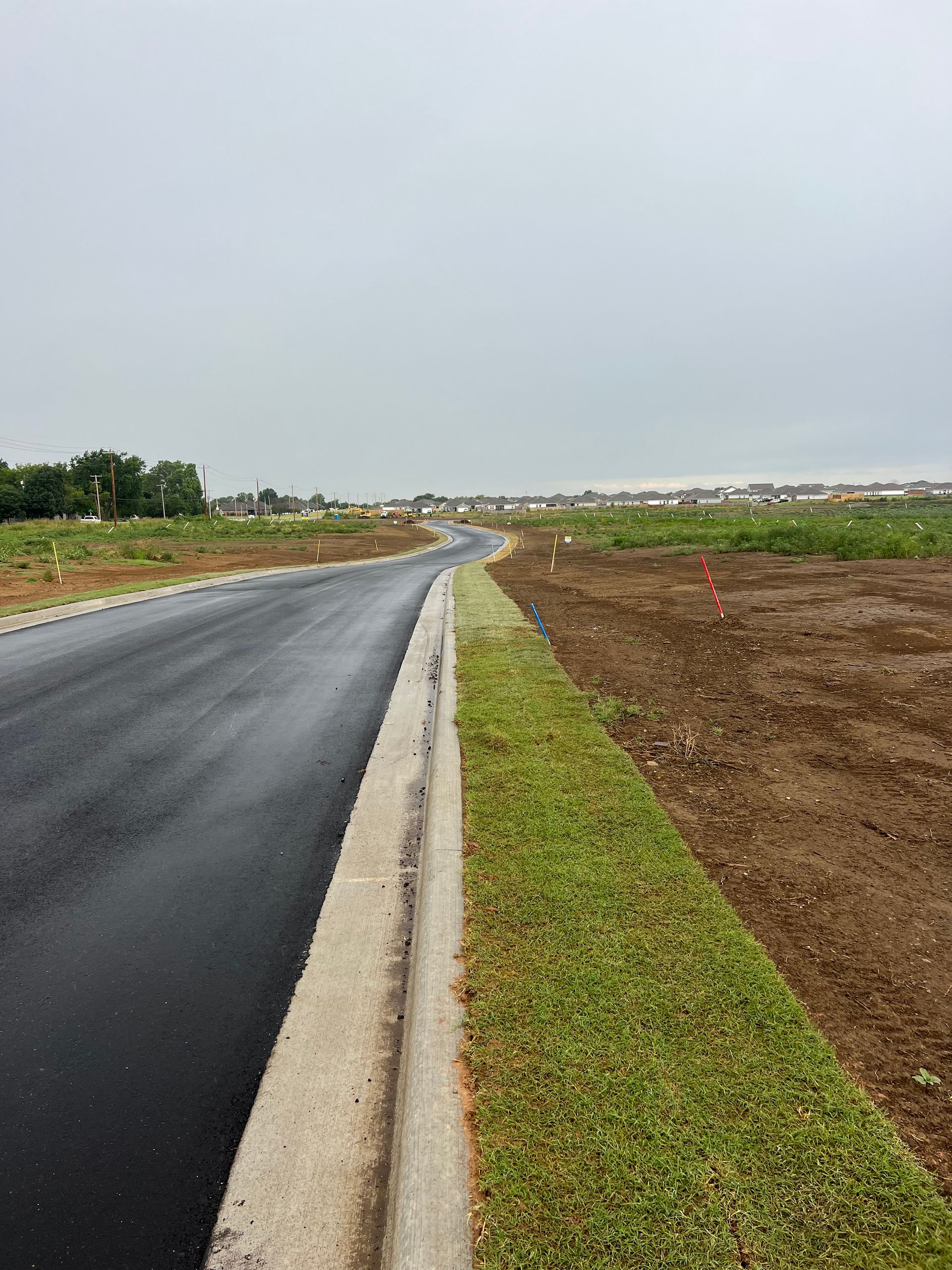 Newly paved road curving through a grassy verge and open land; houses in the background under a cloudy sky.