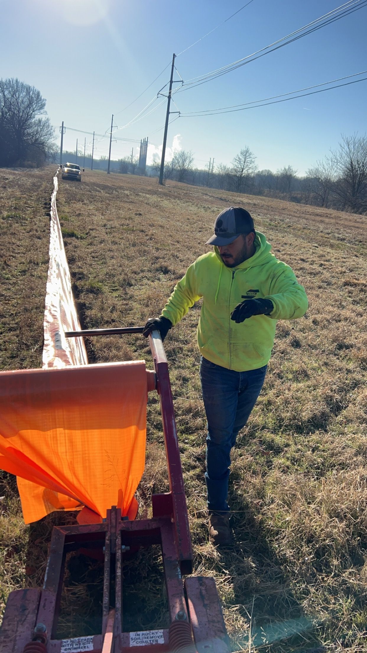 Man in neon yellow jacket, pushing a red cart with orange fabric on a grassy hillside, power lines in background.