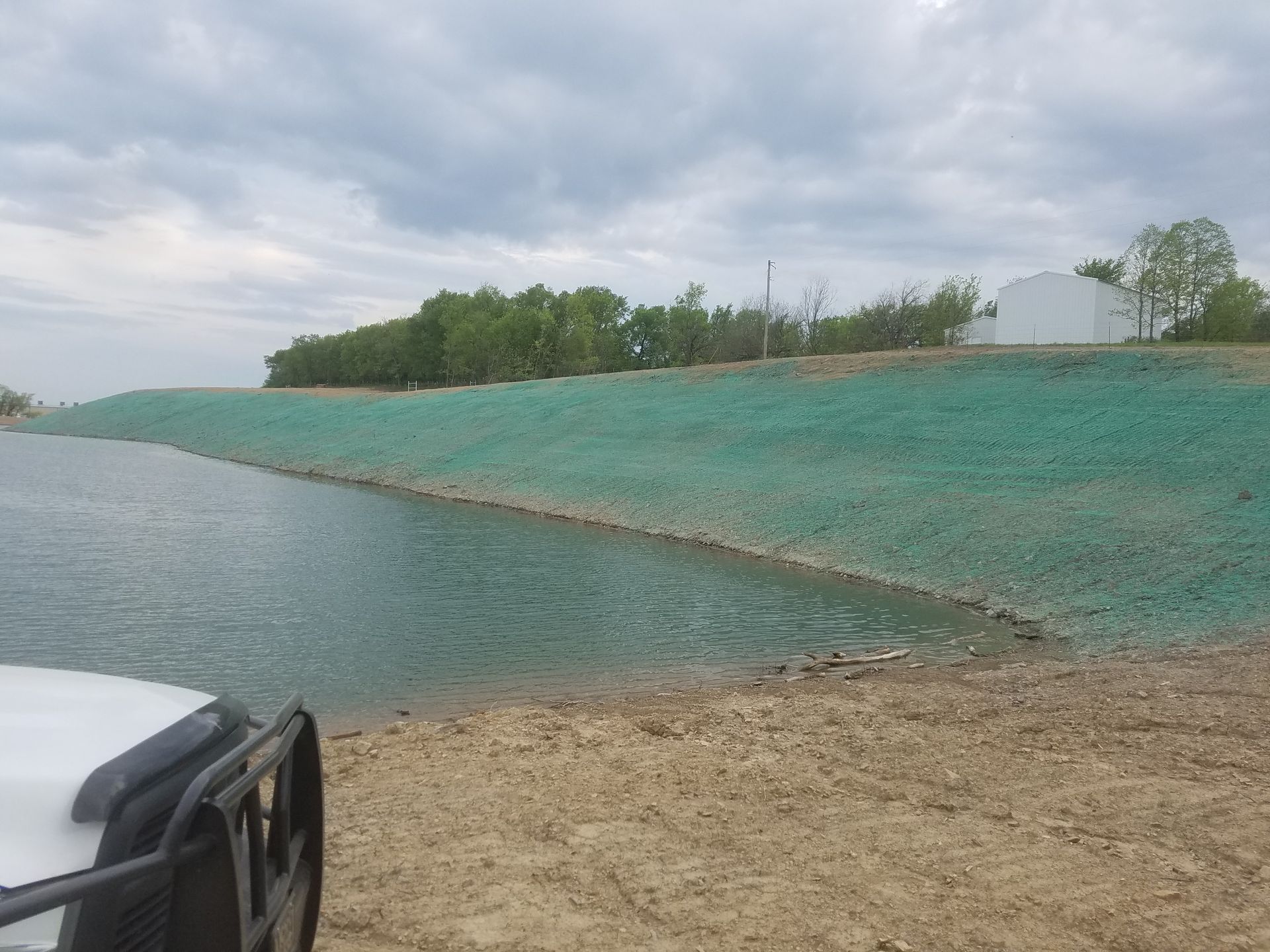 Green-covered slope next to water, possibly for erosion control. Cloudy sky, trees and a white building in the background.