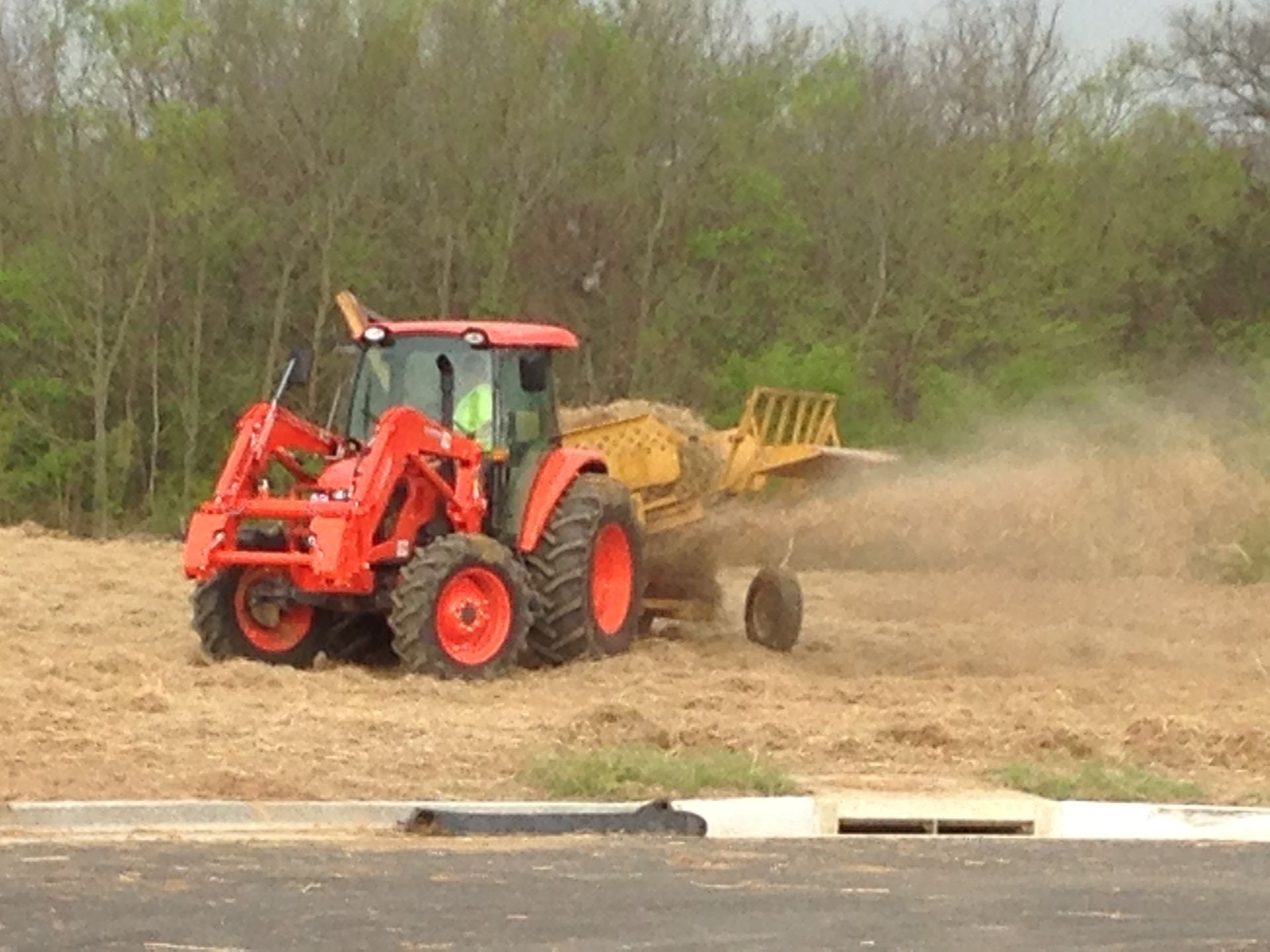 Orange tractor rakes hay in a field near trees under a cloudy sky.