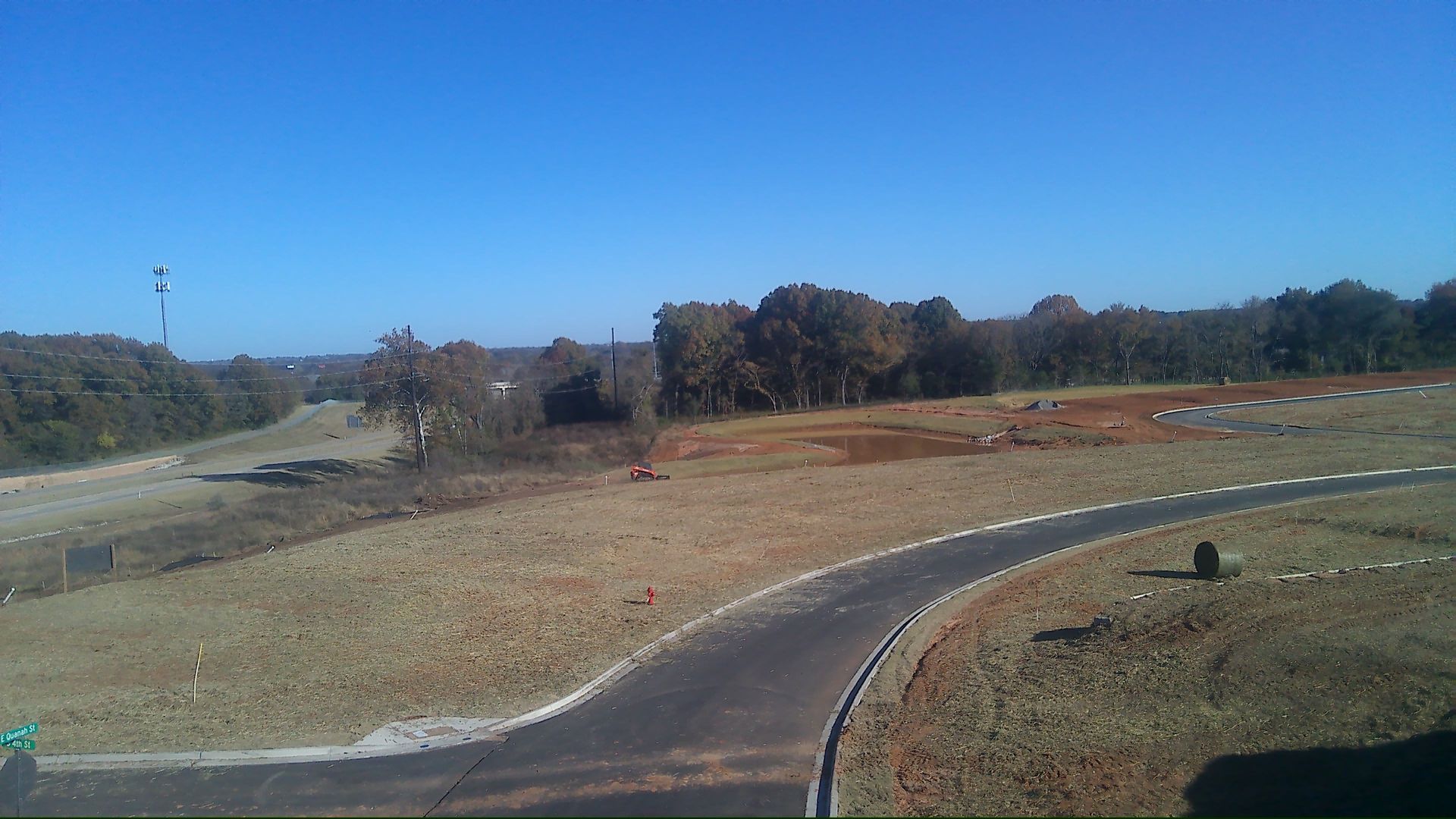 A paved road curves through a brown field under a clear blue sky, with trees in the background.