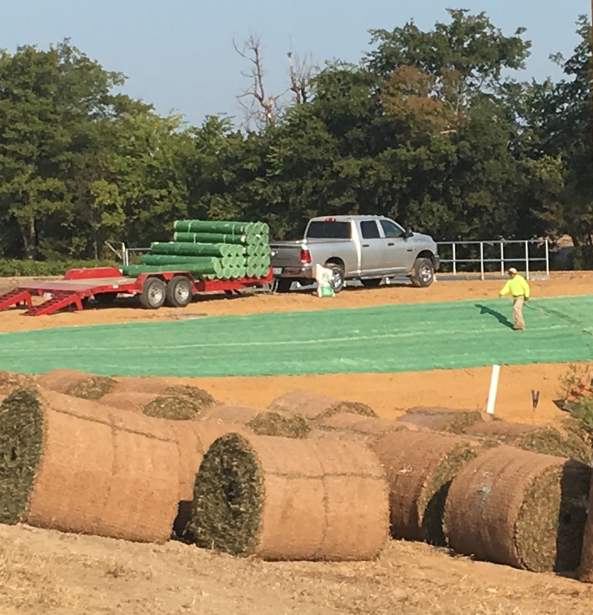 A truck with a trailer carrying rolls of sod. A person is walking on a field of green.