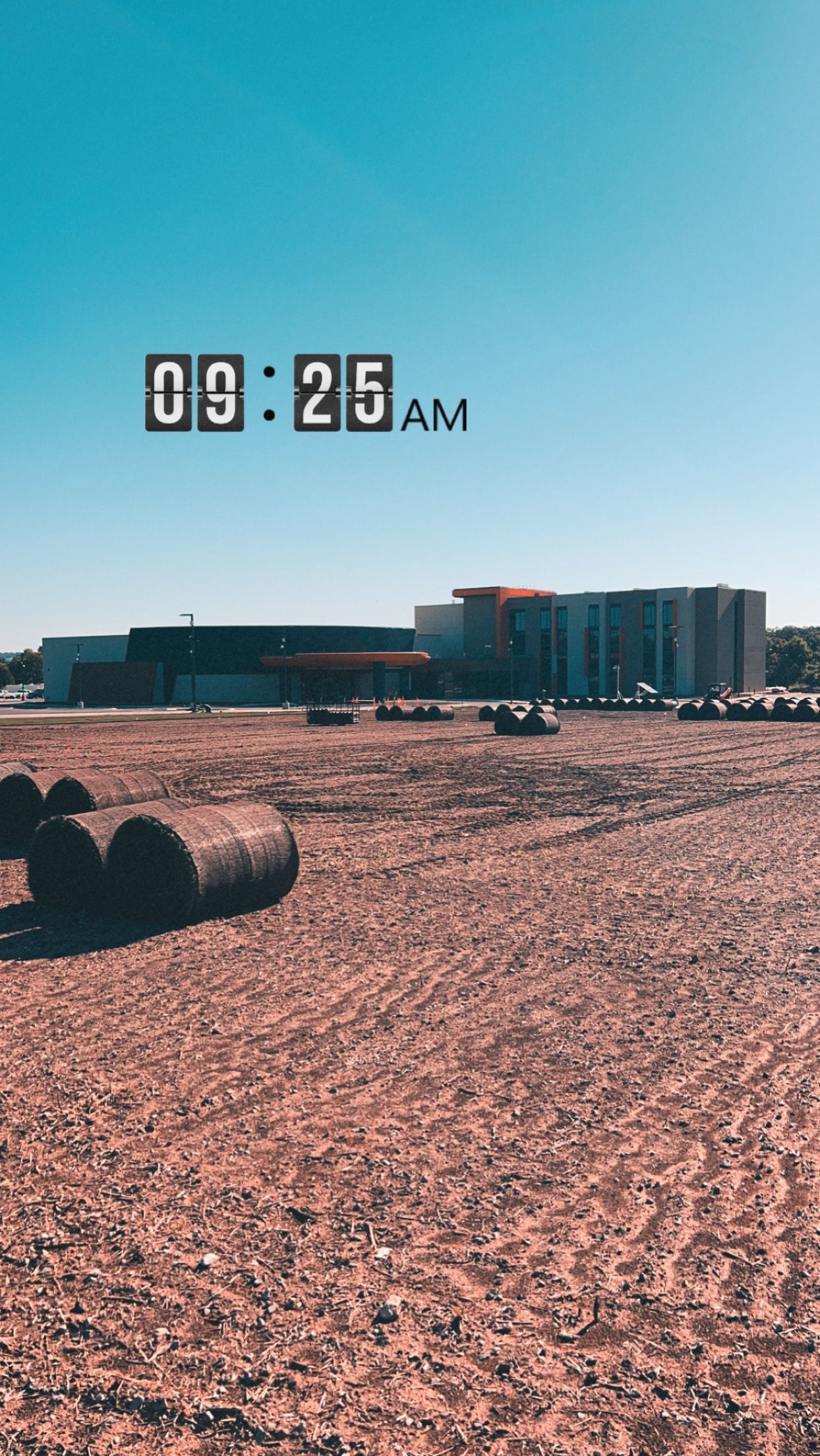 Hay bales in a field, a modern building under a clear blue sky, time stamp: 09:25 AM.