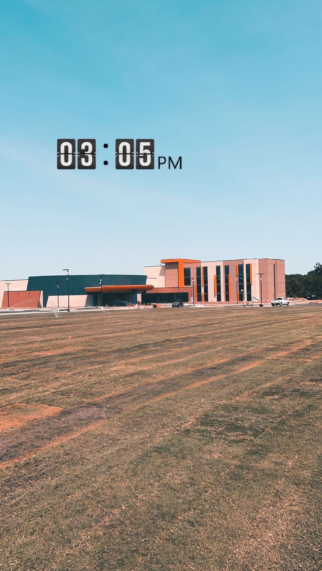 A building with a brick facade in a field under a blue sky, with the time 03:05 PM displayed.