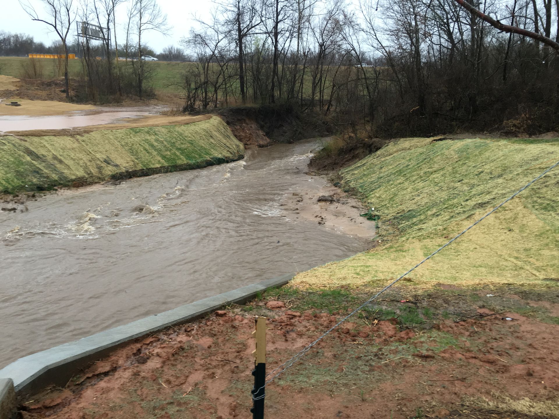 Muddy creek flowing through a channel with grass-covered banks, trees in background.