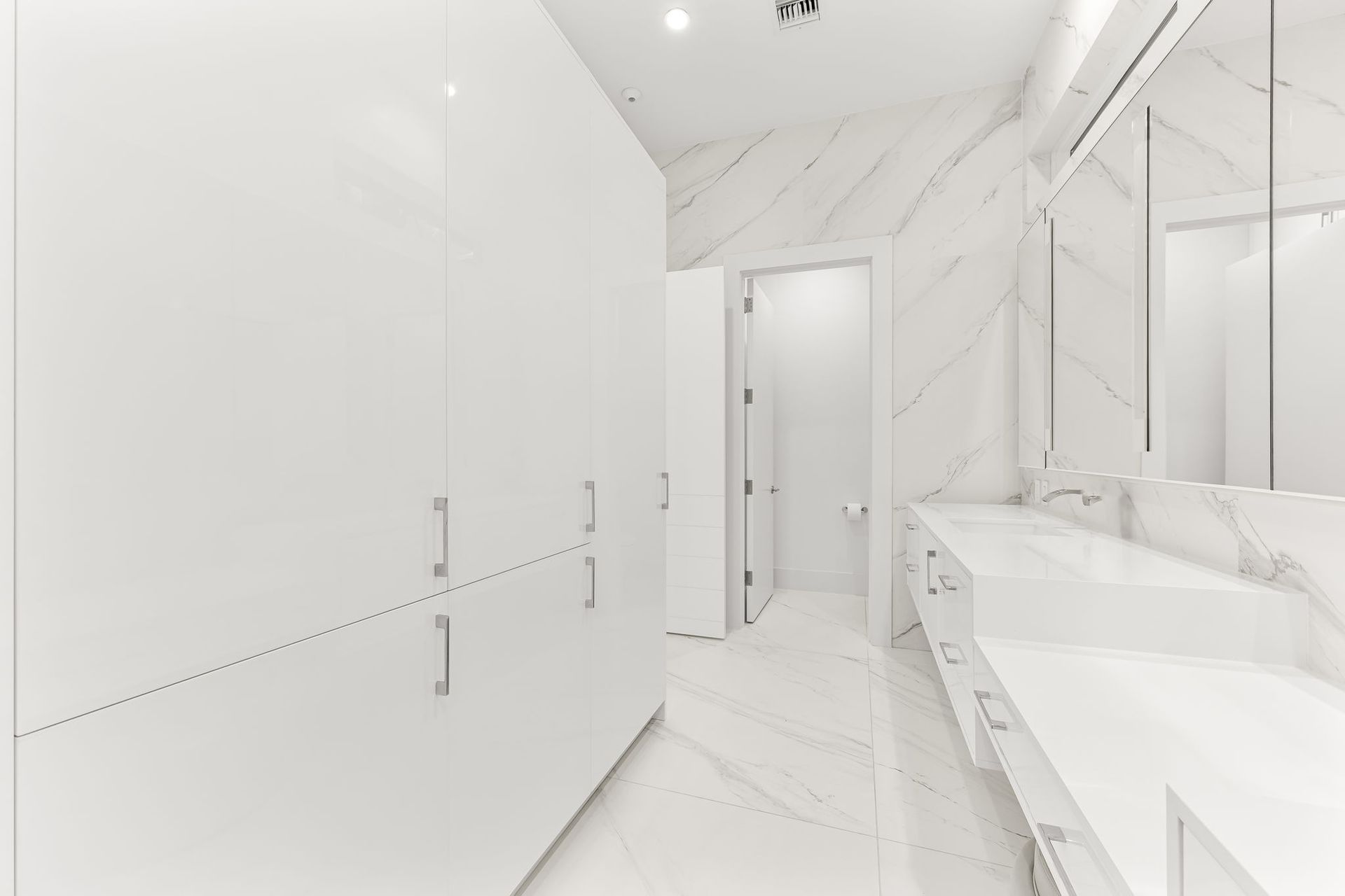White bathroom with marble walls and floor, long vanity, and tall cabinets.