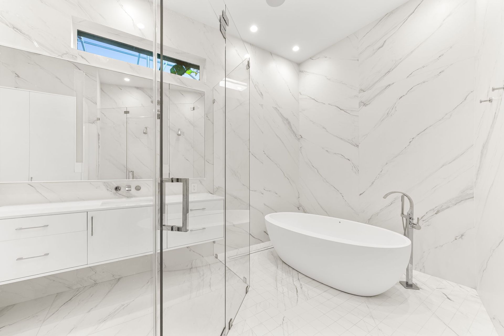 Modern white bathroom with marble walls, glass shower, and oval bathtub.