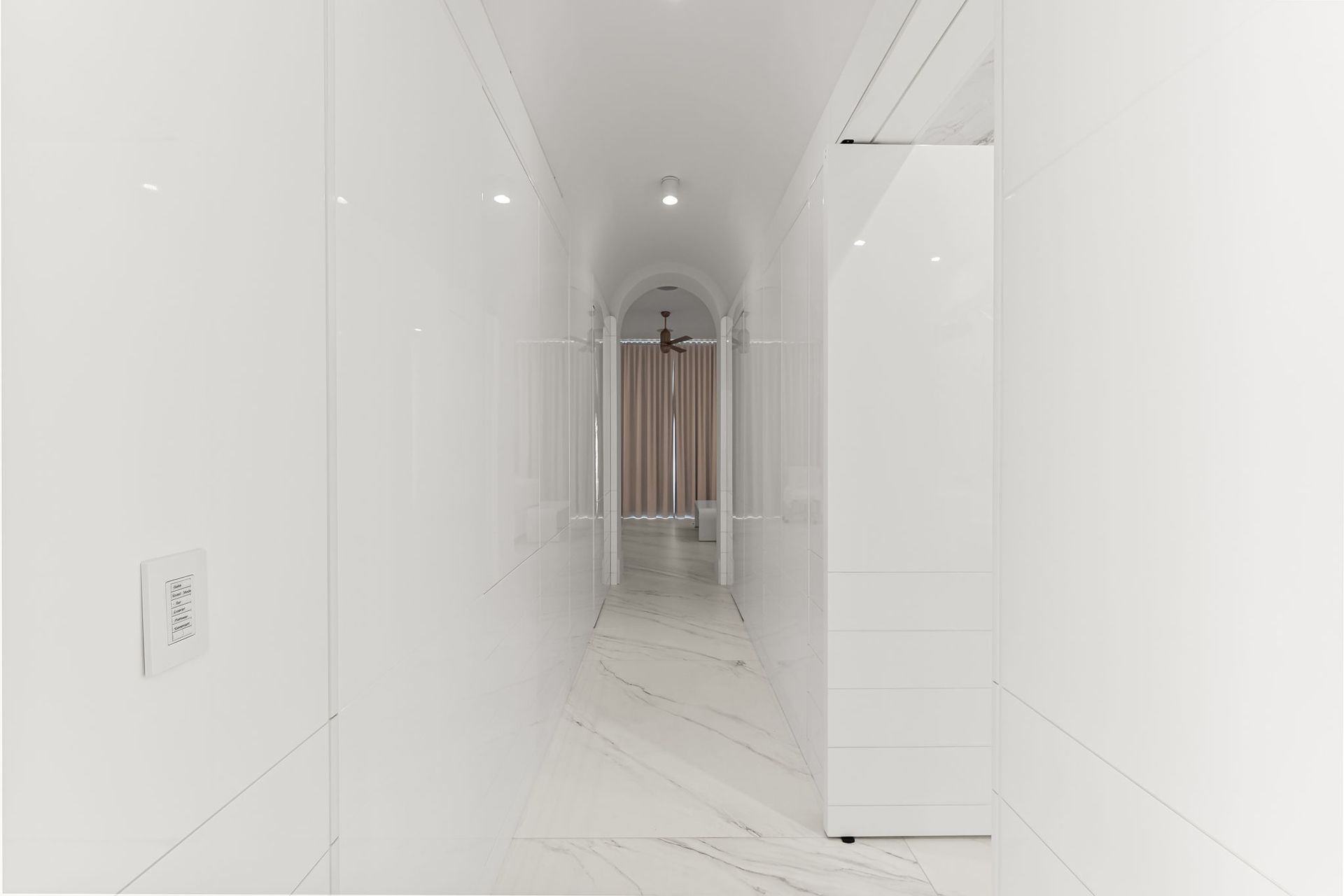 Long, narrow white hallway with archway leading to a room with tan curtains.