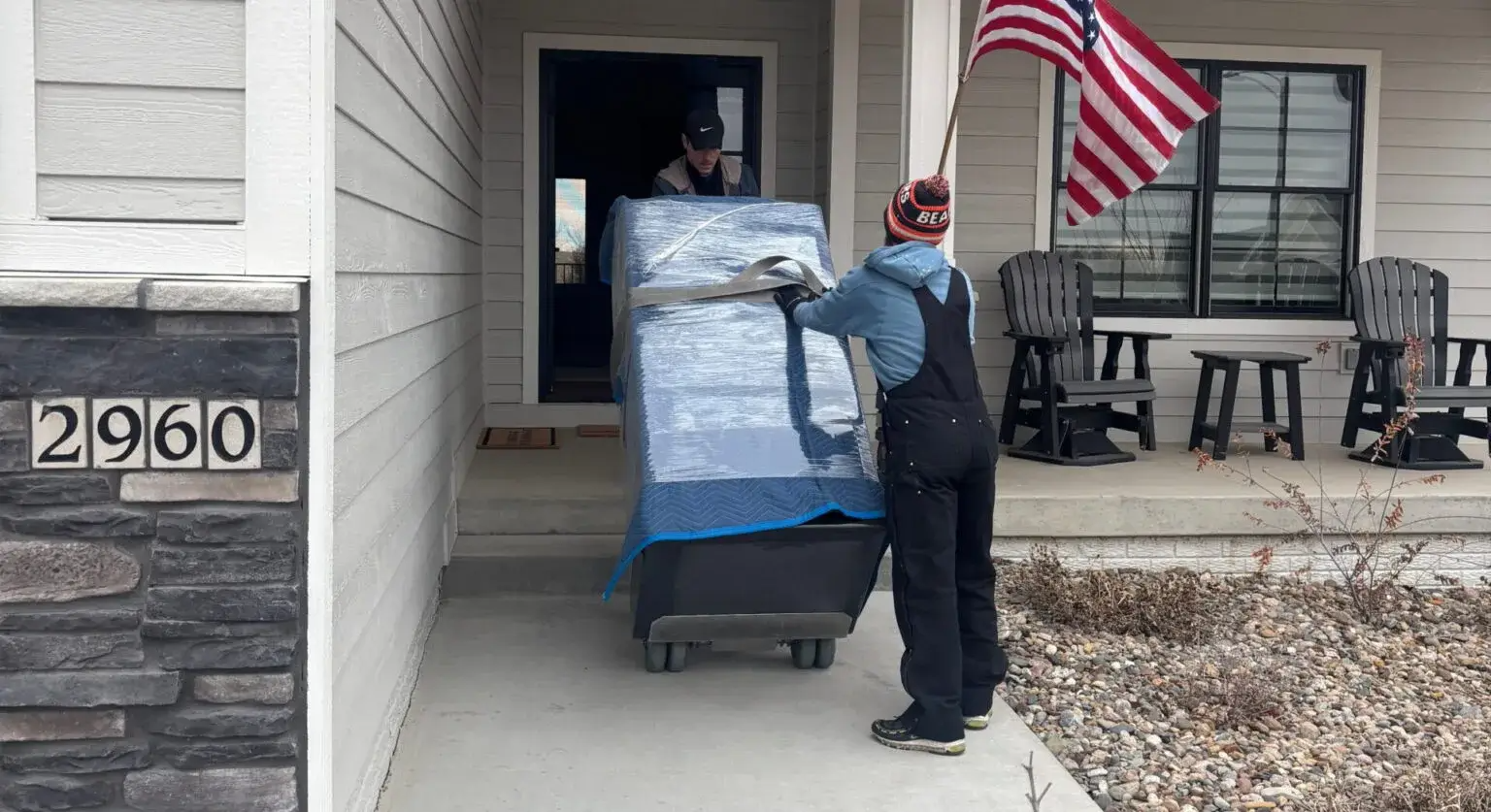 Person standing outside a house beside a dolly loaded with wrapped furniture and a U.S. flag
